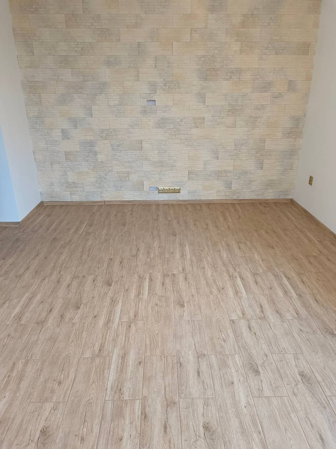 Wnętrze pokoju po remoncie: ułożone panele podłogowe w odcieniach jasnego drewna i ściana wykończona dekoracyjną płytką imitującą kamień w beżowo-szarej kolorystyce, widoczne gniazdka elektryczne.