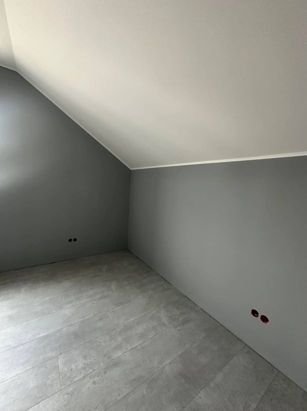 Pomalowane na szaro poddasze ze świeżo ułożonymi panelami podłogowymi imitującymi beton, widoczne gniazdka elektryczne bez osprzętu.