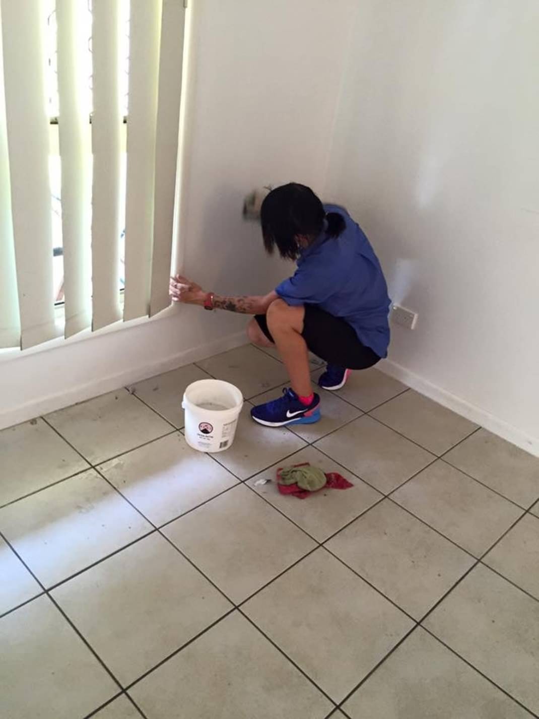 Osoba w niebieskiej koszuli i sportowych butach maluje białą ścianę w rogu pokoju, obok okna z żaluzjami, widoczne wiadro z farbą i szmatka na kafelkach podłogowych.