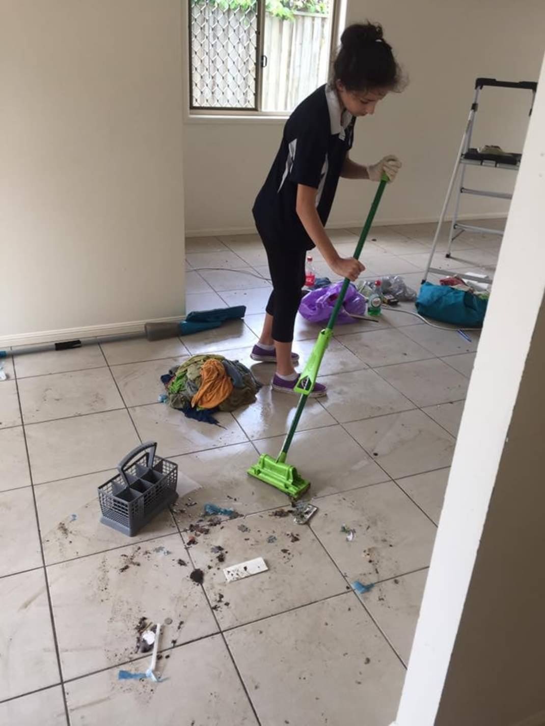 Młoda kobieta w stroju roboczym, z gumowymi rękawicami, mopem na długim kiju i fioletowych tenisów na nogach, sprząta zabrudzoną podłogę z płytek w pustym pokoju, gdzie w tle widoczna jest drabina.