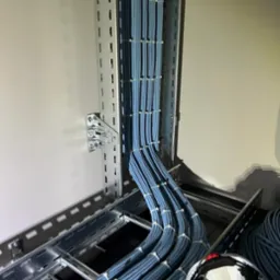 Równolegle ułożone, niebieskie przewody w metalowej korycie kablowej, zabezpieczone opaskami. Widoczny fragment kasku ochronnego.