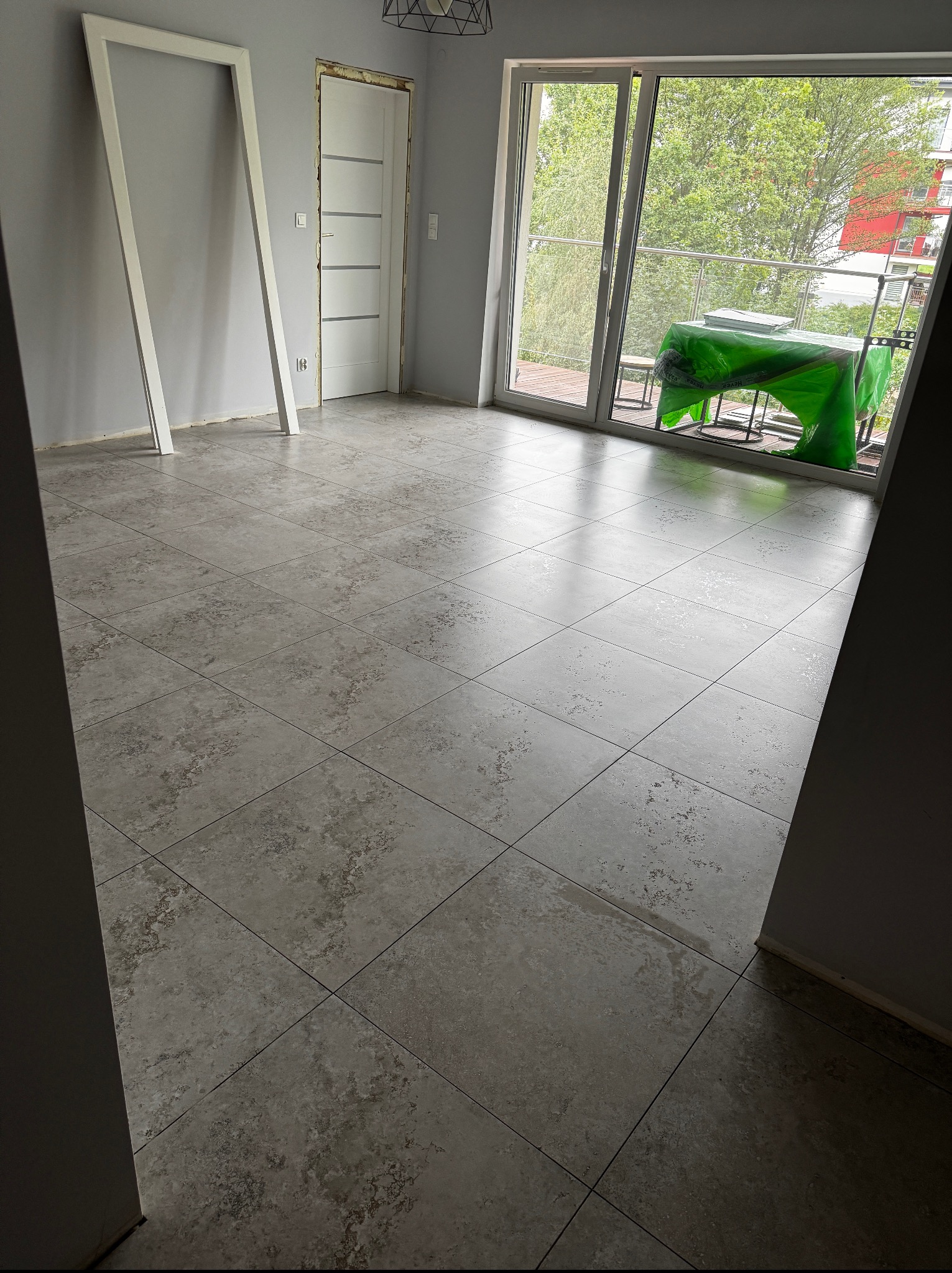 Wykończona podłoga z szarych płytek w jasnym pokoju z oknem balkonowym, widoczne przygotowania do dalszych prac, np. ościeżnica oparta o ścianę, drzwi.