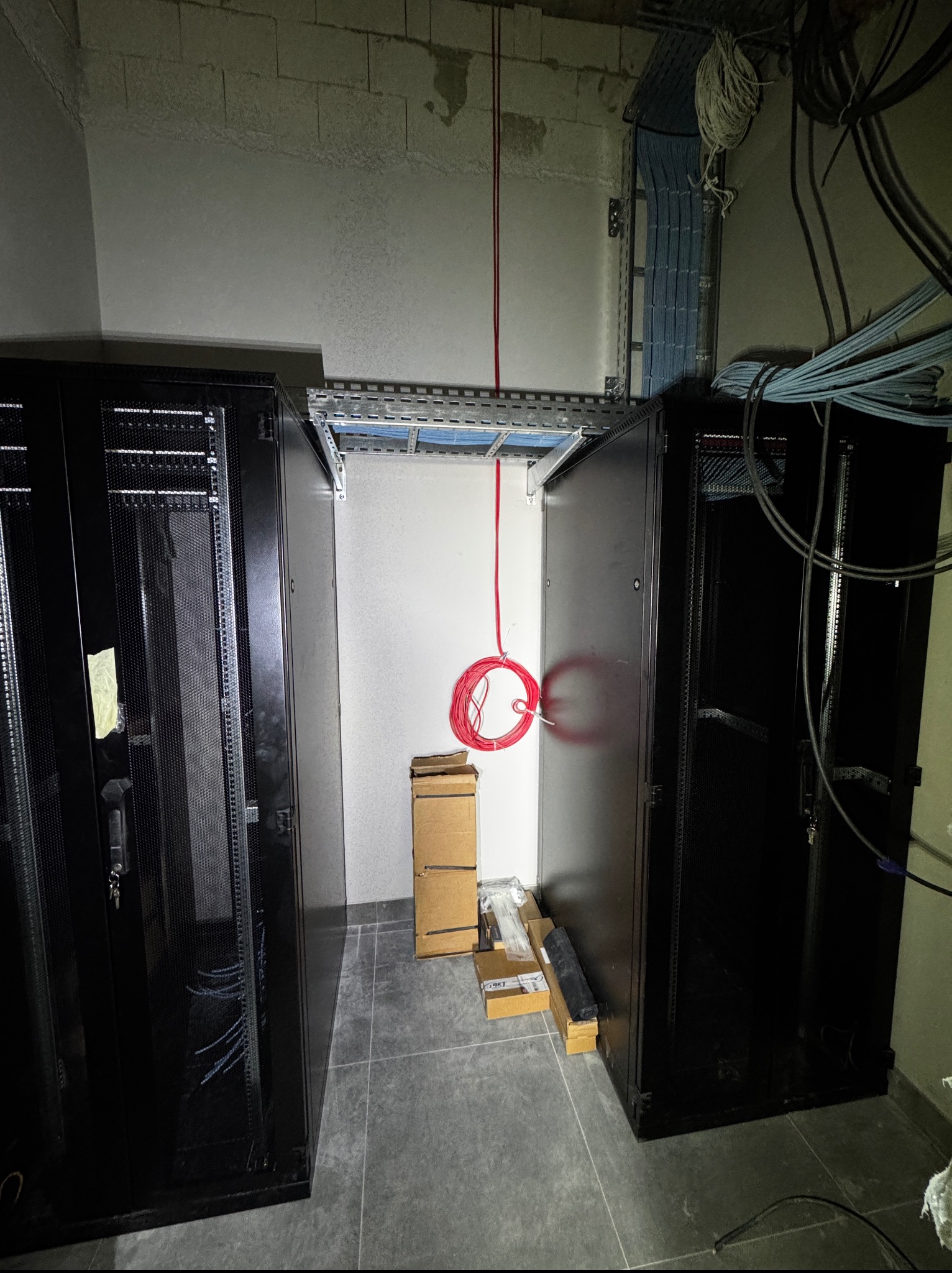 Wnętrze serwerowni w trakcie budowy: szafy rack, kable, koryta kablowe pod sufitem, betonowy sufit, podłoga z płytek. Widoczne przygotowania do instalacji.