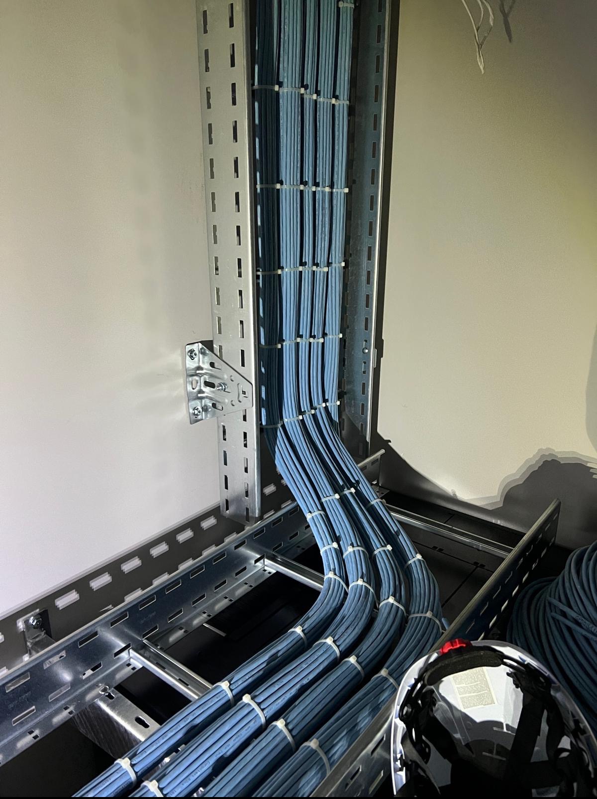 Równolegle ułożone, niebieskie przewody w metalowej korycie kablowej, zabezpieczone opaskami. Widoczny fragment kasku ochronnego.