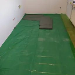 Przygotowanie do układania paneli: rozłożona folia ochronna na podłodze, płyty podkładowe i wiaderko z drobnymi elementami w rogu pokoju.