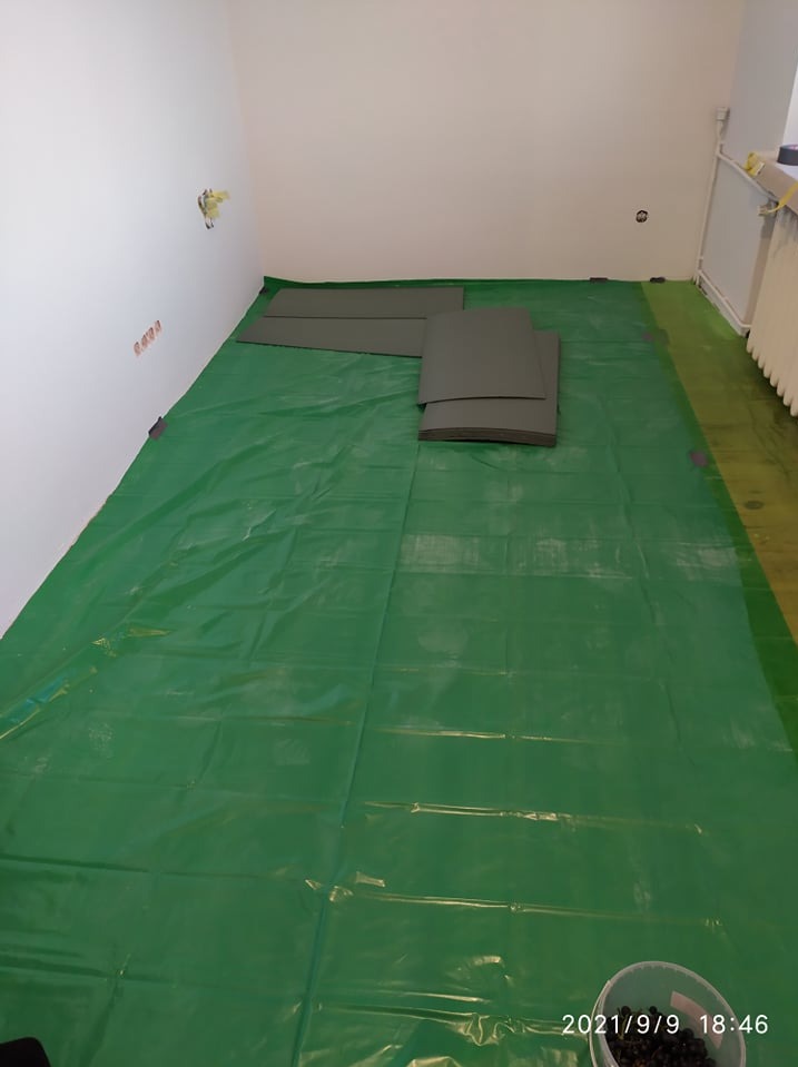 Przygotowanie do układania paneli: rozłożona folia ochronna na podłodze, płyty podkładowe i wiaderko z drobnymi elementami w rogu pokoju.