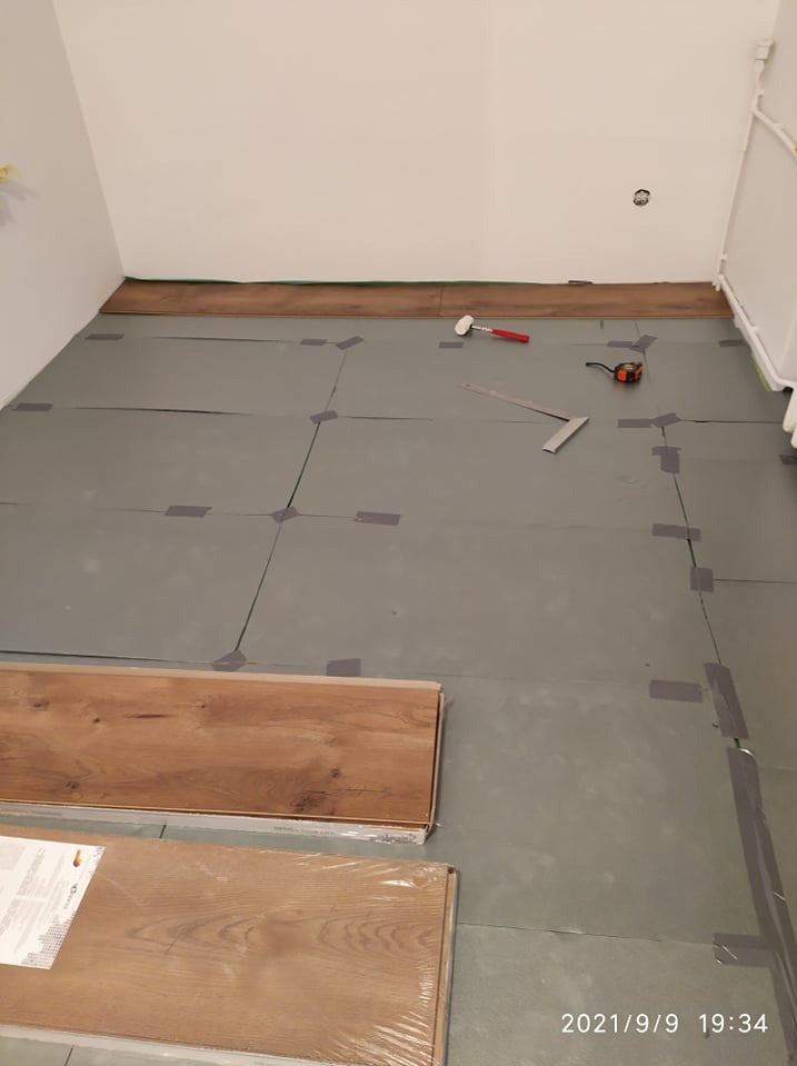 Przygotowanie do montażu paneli podłogowych: rozłożona izolacja z zaznaczonymi liniami cięcia i narzędzia - młotek, kątownik, miarka. Na pierwszym planie paczki paneli w folii.