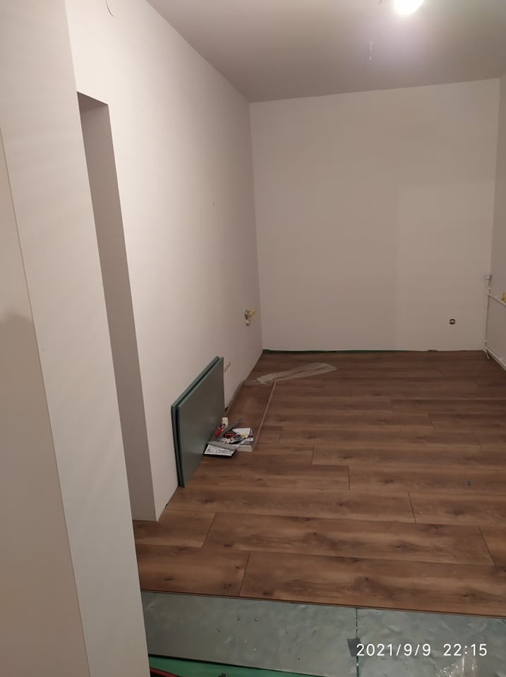 Fragment pokoju z częściowo ułożonymi panelami podłogowymi w odcieniach brązu; widoczne narzędzia i paczka paneli oparta o ścianę; podłoże pod panele częściowo odsłonięte.