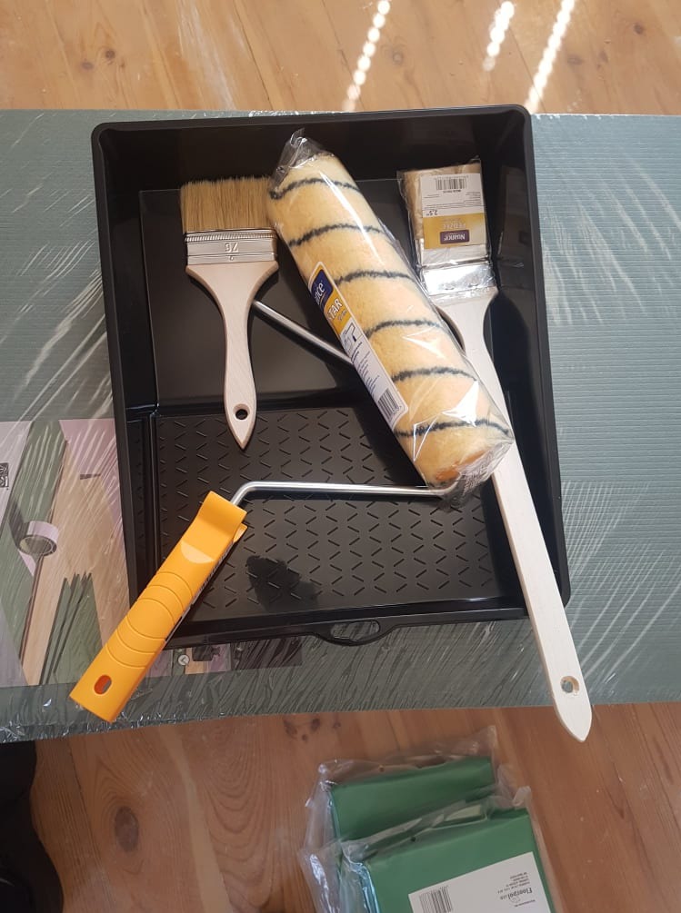 Zestaw malarski w czarnej kuwecie: pędzel 76 mm, wałek z czarno-żółtymi paskami w folii, mały pędzel w folii i uchwyt do wałka z żółtą rączką, na tle zabezpieczonej folią podłogi.