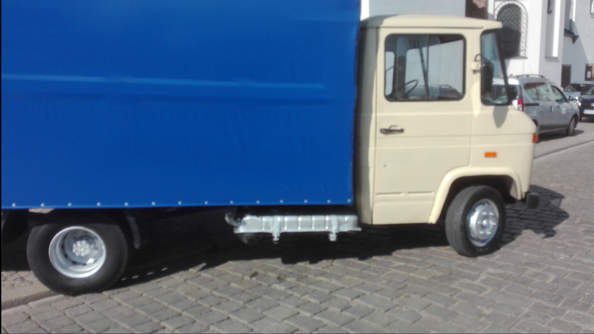 Beżowy samochód dostawczy z niebieską plandeką na kostce brukowej, widok z boku.