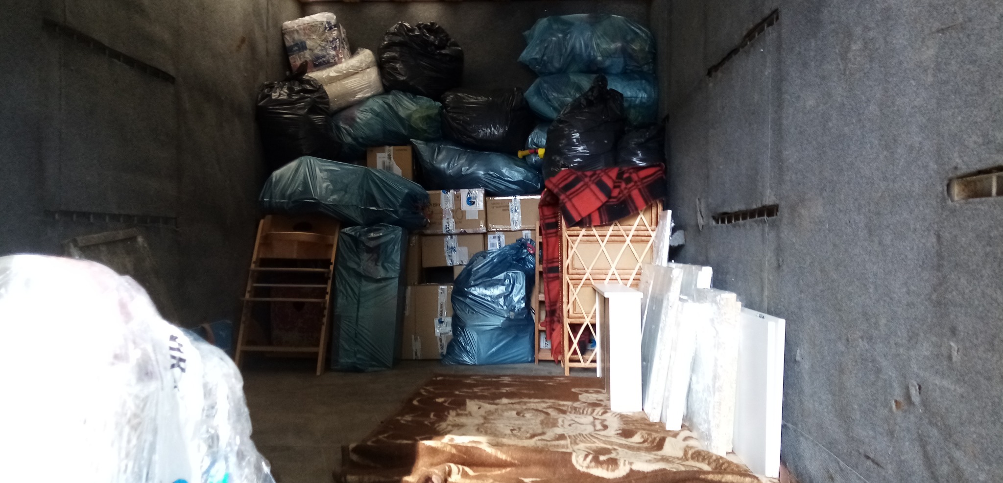 Wnętrze przestrzeni ładunkowej wypełnione kartonami, workami z zawartością, drewnianymi meblami i panelami, zabezpieczonymi folią, ułożonymi warstwami, widoczny dywan na podłodze.