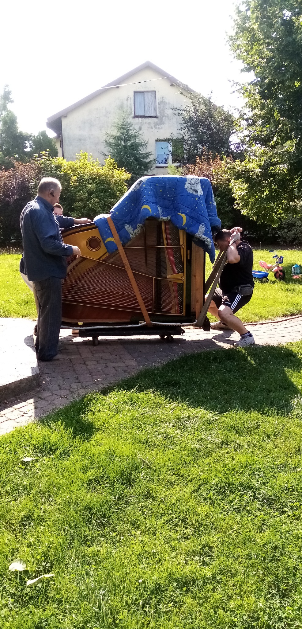 Dwóch mężczyzn przenosi pianino na wózku, zabezpieczone pasami i kocem w niebieskie gwiazdki, na tle trawnika i budynku.