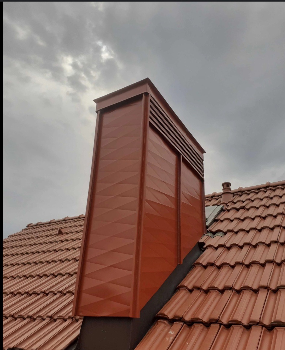 Nowo pomalowany, brązowy komin z dekoracyjnym wzorem na tle pochmurnego nieba, widoczny na tle czerwonej dachówki.