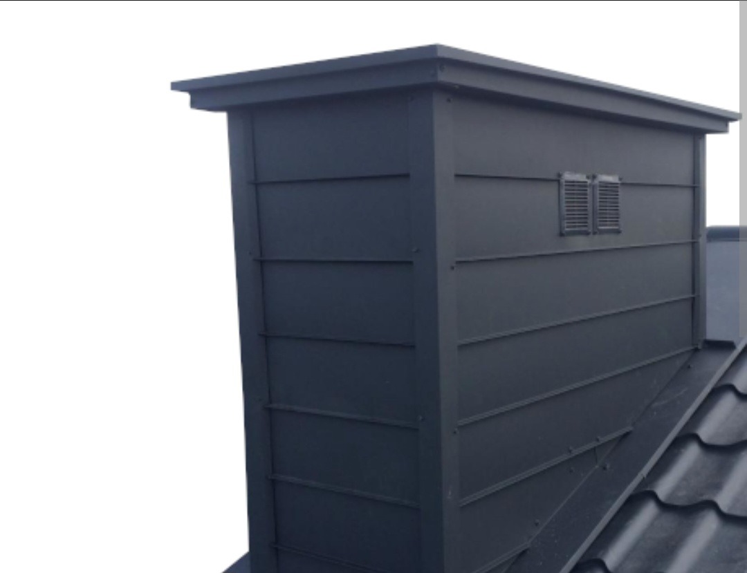 Metalowy komin wentylacyjny w kolorze antracytowym, zamontowany na dachu pokrytym ciemną dachówką, z widocznymi kratkami wentylacyjnymi.