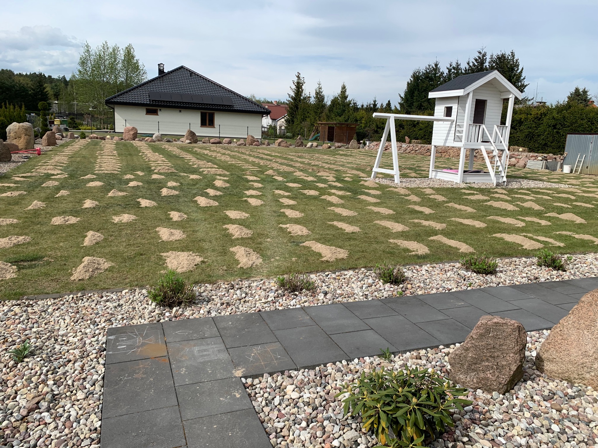 Przygotowany teren pod trawnik z nasypanymi kopczykami ziemi, widoczny domek dla dzieci i dom jednorodzinny w tle.