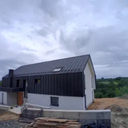 Nowoczesny dom jednorodzinny z czarnym dachem z blachy na rąbek stojący i jasną elewacją, w trakcie prac wykończeniowych na zewnątrz, widoczne materiały budowlane i krajobraz w tle.