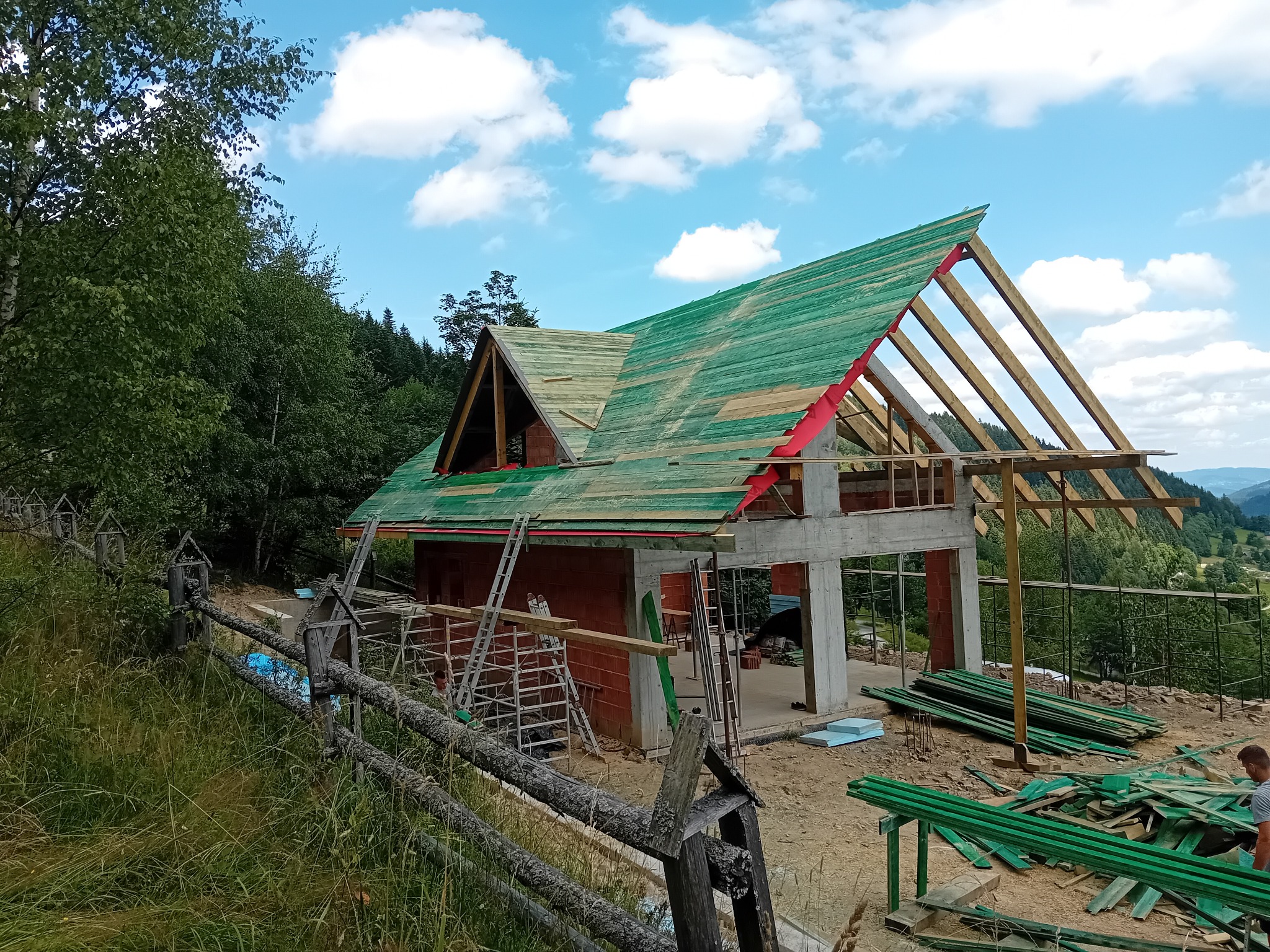 Budowa dachu: widok na konstrukcję więźby dachowej z deskowaniem pokrytym zieloną folią, częściowo odkrytą, z elementami czerwonej membrany, na tle murowanego domu w stanie surowym otoczonego...