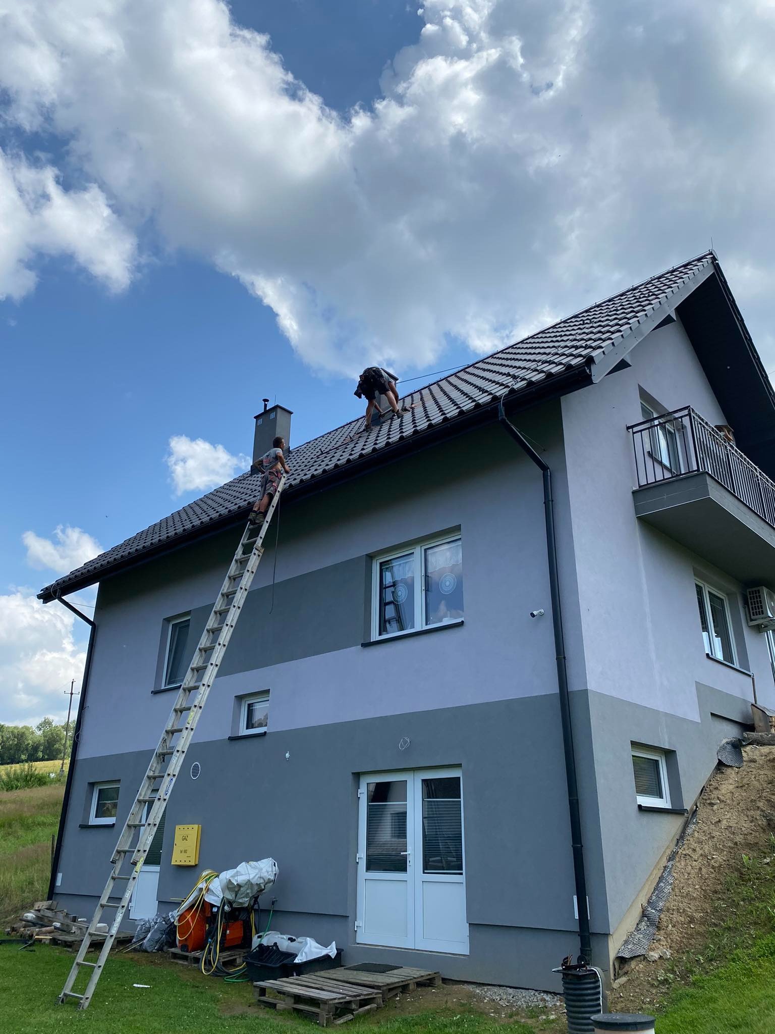 Pracownicy na dachu dwukondygnacyjnego domu z szarej elewacją i balkonem, jeden na drabinie, drugi na dachu, błękitne niebo z chmurami w tle.