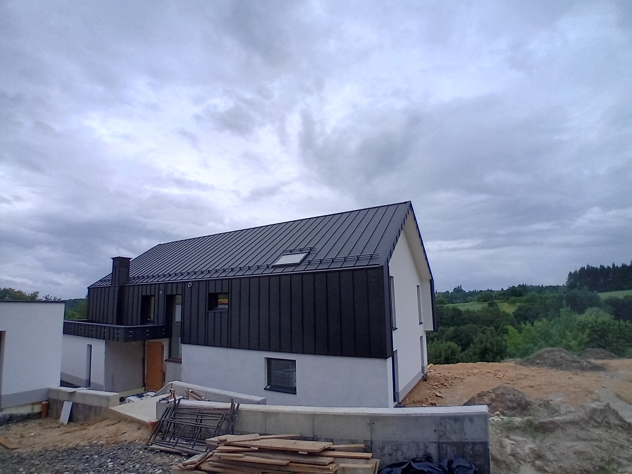 Nowoczesny dom jednorodzinny z czarnym dachem z blachy na rąbek stojący i jasną elewacją, w trakcie prac wykończeniowych na zewnątrz, widoczne materiały budowlane i krajobraz w tle.