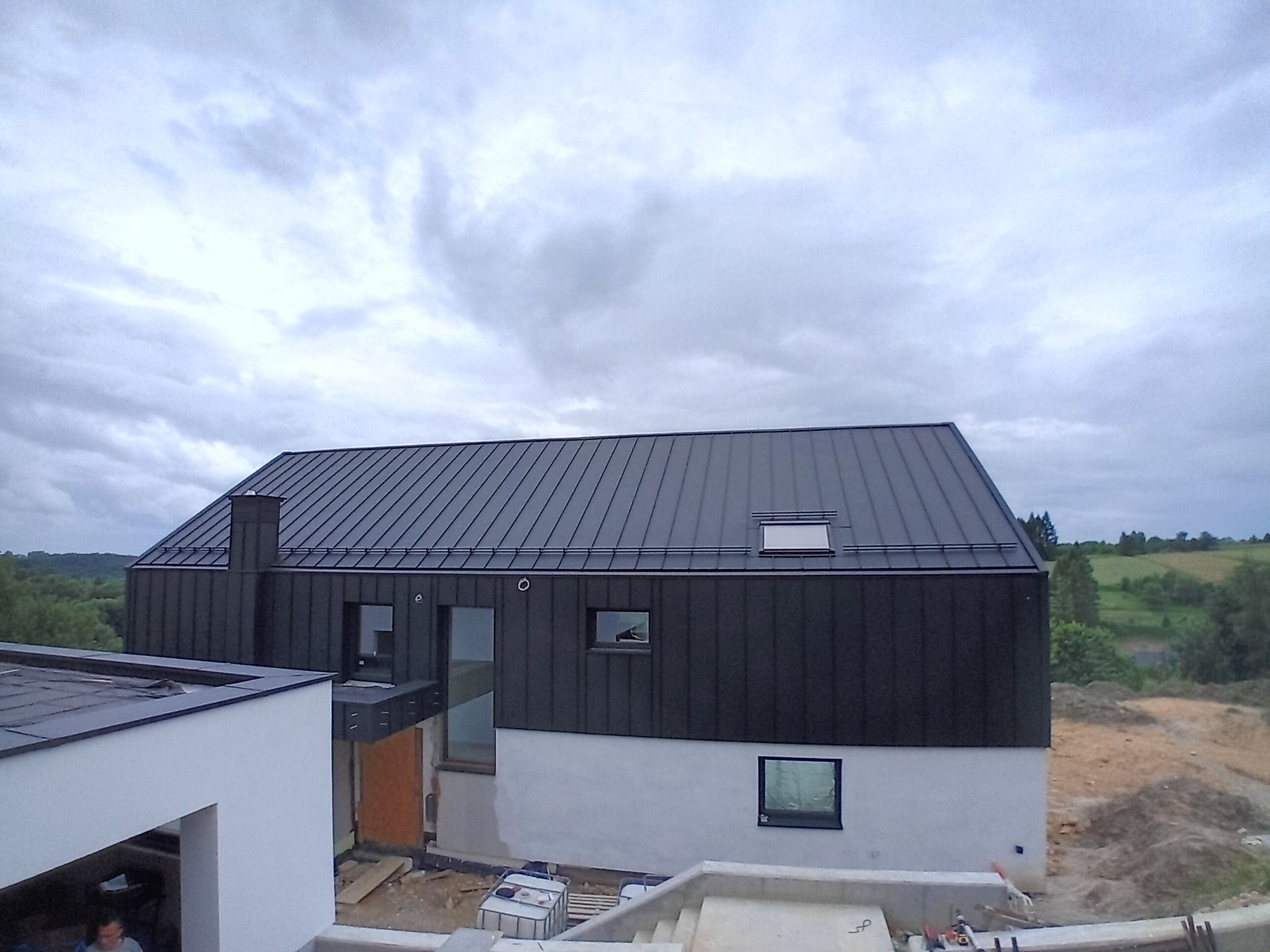 Dom jednorodzinny z ciemną, metalową obróbką dachu i elewacją częściowo wykończoną na biało, widok z góry na tle pochmurnego nieba i zieleni w oddali.