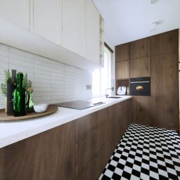 Kreska Karolina Kuźmicka - Nowoczesna kuchnia z białymi szafkami górnymi i drewnianymi dolnymi, czarno-biała szachownica na podłodze, dekoracyjne butelki i zioła na blacie, piekarnik z pieczonym kurczakiem w tle.