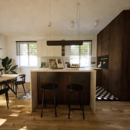 Kreska Karolina Kuźmicka - Wnętrze kuchni z wyspą, drewnianymi szafkami i podłogą, czarno-biała szachownica na podłodze w głębi, książka kucharska i owoce na blacie wyspy.