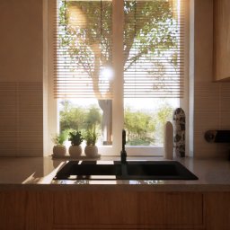 Kreska Karolina Kuźmicka - Nowoczesna kuchnia z drewnianymi szafkami, czarnym zlewem i baterią, oraz oknem z żaluzjami, przez które wpada naturalne światło, oświetlając rośliny w doniczkach na parapecie.