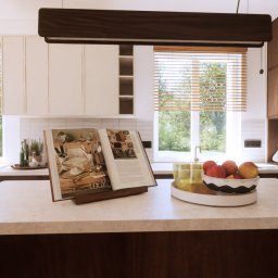 Kreska Karolina Kuźmicka - Wnętrze kuchni z drewnianymi i białymi szafkami, oświetleniem podszafkowym i oknami z żaluzjami. Na blacie otwarta książka kucharska i miska z owocami.