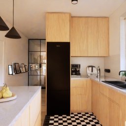 Kreska Karolina Kuźmicka - Jasna kuchnia z drewnianymi szafkami, czarnym zlewem i armaturą, czarną lodówką Bosch i podłogą w szachownicę; na blacie leżą dekoracyjne gruszki, a nad wyspą kuchenną wiszą trzy czarne lampy...