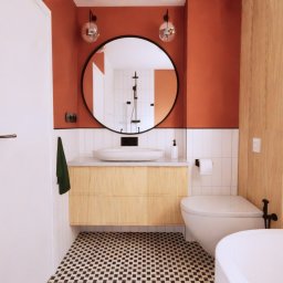 Kreska Karolina Kuźmicka - Nowoczesna łazienka z terakotową ścianą, okrągłym lustrem z czarną ramą, białą umywalką nablatową na szafce z jasnego drewna, toaletą i fragmentem białej wanny; podłoga w czarno-białą szachownicę.