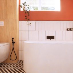 Kreska Karolina Kuźmicka - Nowoczesna łazienka z wiszącą toaletą, bidetką i wolnostojącą wanną na tle terakotowej ściany i białych płytek, podkreślona czarno-białą szachownicą na podłodze i wiszącą rośliną przy oknie.