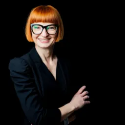 Portret uśmiechniętej kobiety w czarnej marynarce i okularach na czarnym tle, z rudymi włosami z grzywką.