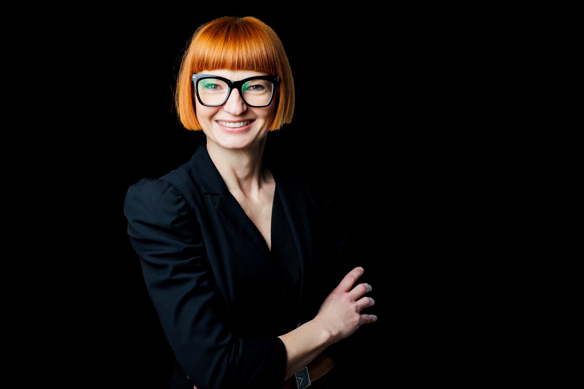 Portret uśmiechniętej kobiety w czarnej marynarce i okularach na czarnym tle, z rudymi włosami z grzywką.