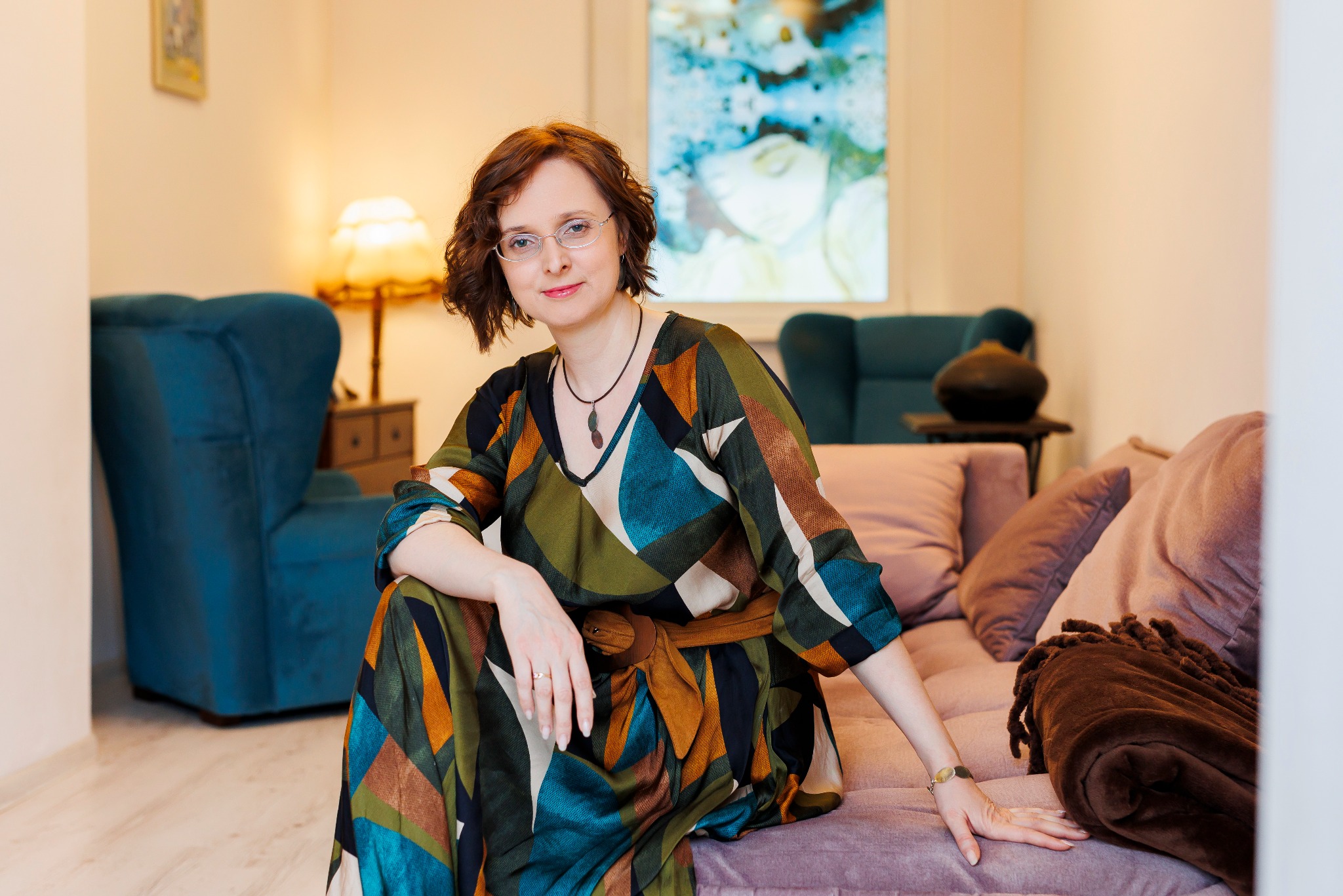 Portret kobiety w okularach, siedzącej na sofie w sukience w geometryczne wzory, z brązowym paskiem i wisiorkiem, w tle niebieski fotel i obraz.