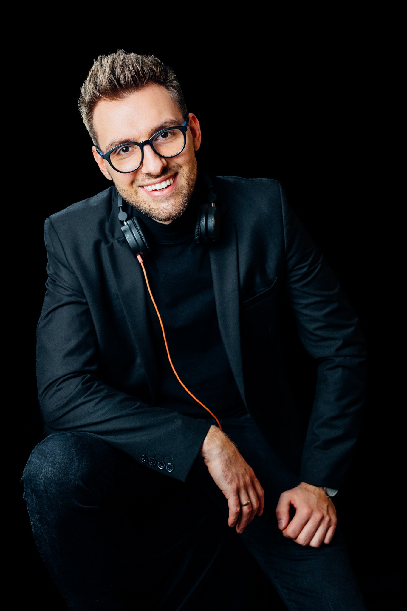 Uśmiechnięty mężczyzna w czarnym garniturze, okularach i słuchawkach na szyi, pozuje na czarnym tle w Rybniku.