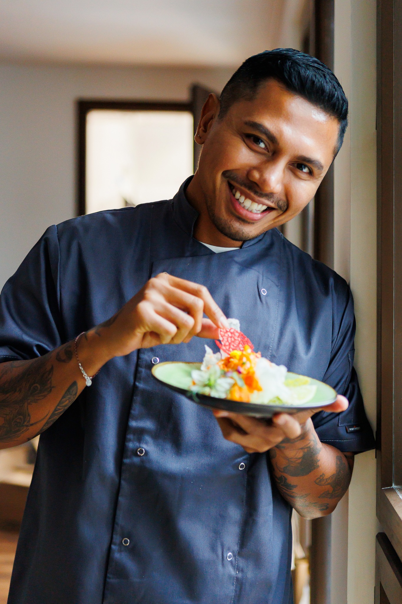Uśmiechnięty kucharz w granatowym uniformie, z tatuażami na rękach, dekoruje talerz z sałatką i plasterkiem limonki czerwoną, ażurową dekoracją.