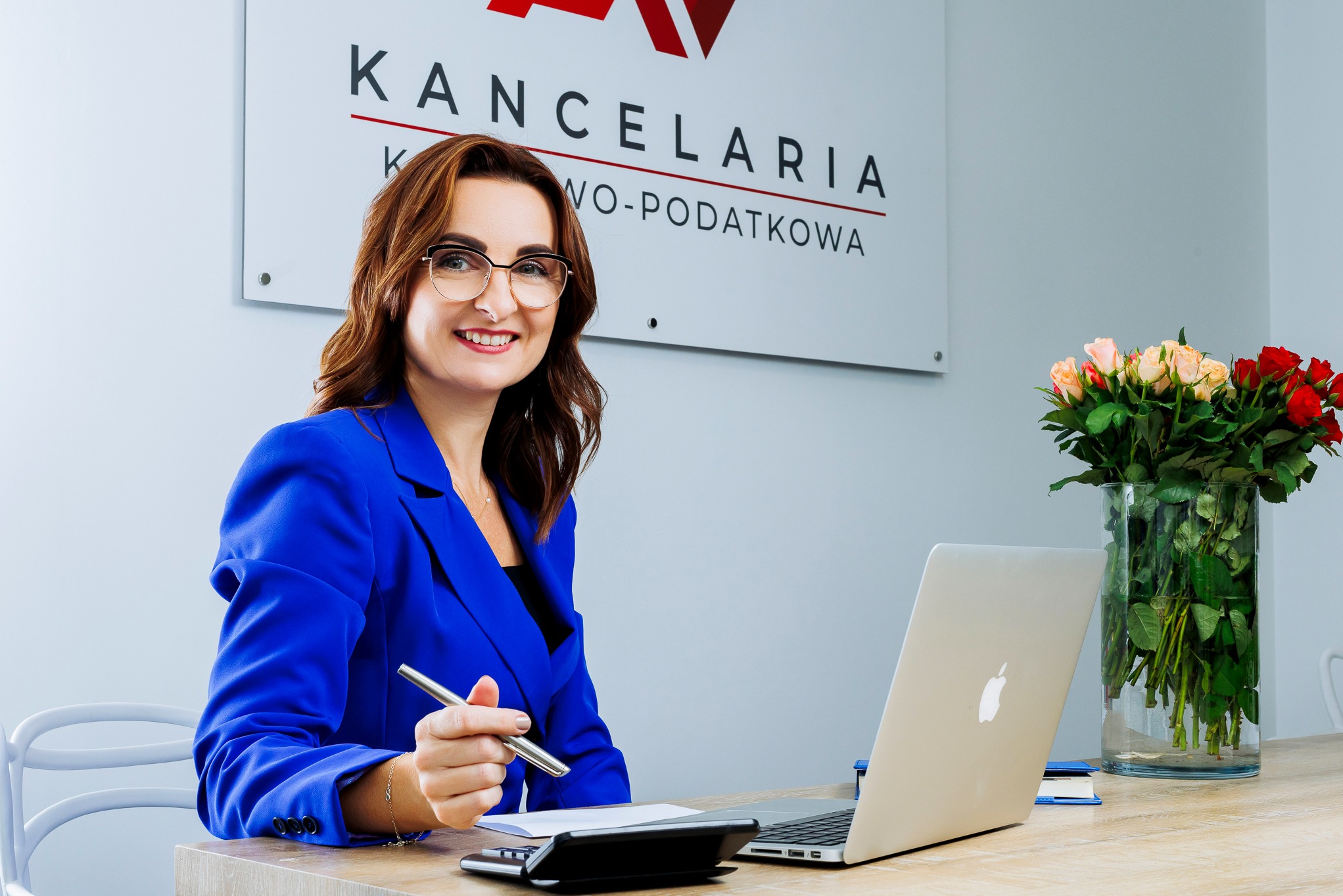 Uśmiechnięta kobieta w niebieskim żakiecie i okularach, siedząca przy biurku z laptopem, trzymająca długopis, z logo kancelarii i wazonem róż w tle.