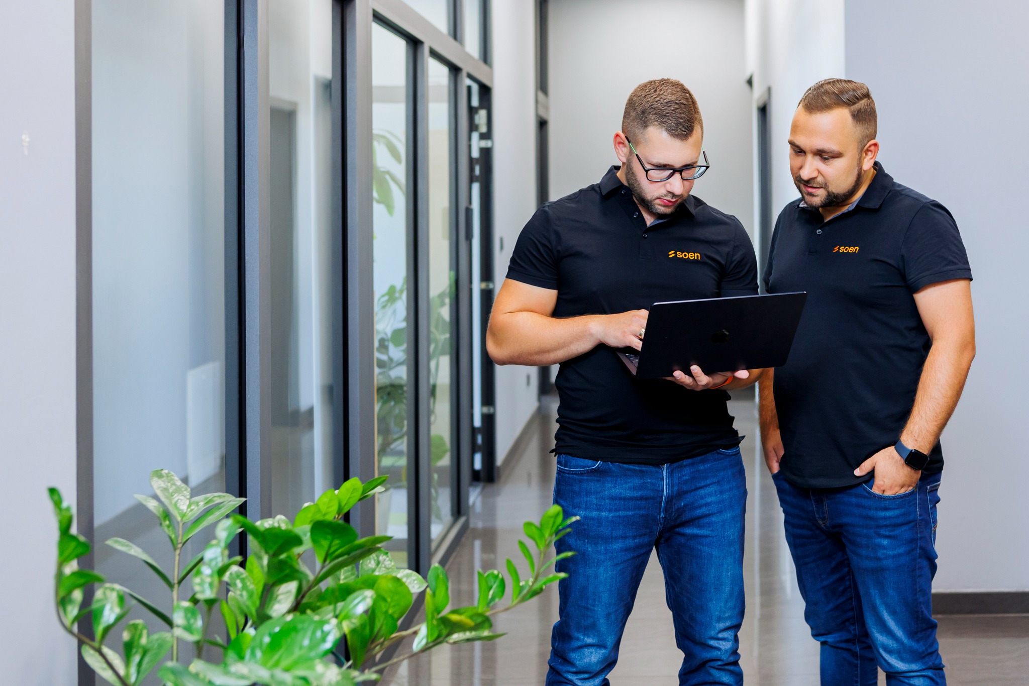 Dwóch mężczyzn w czarnych koszulkach polo z logo firmy, pochylonych nad laptopem w nowoczesnym biurze, z rośliną doniczkową w lewym dolnym rogu i widocznymi przeszkleniami w tle.