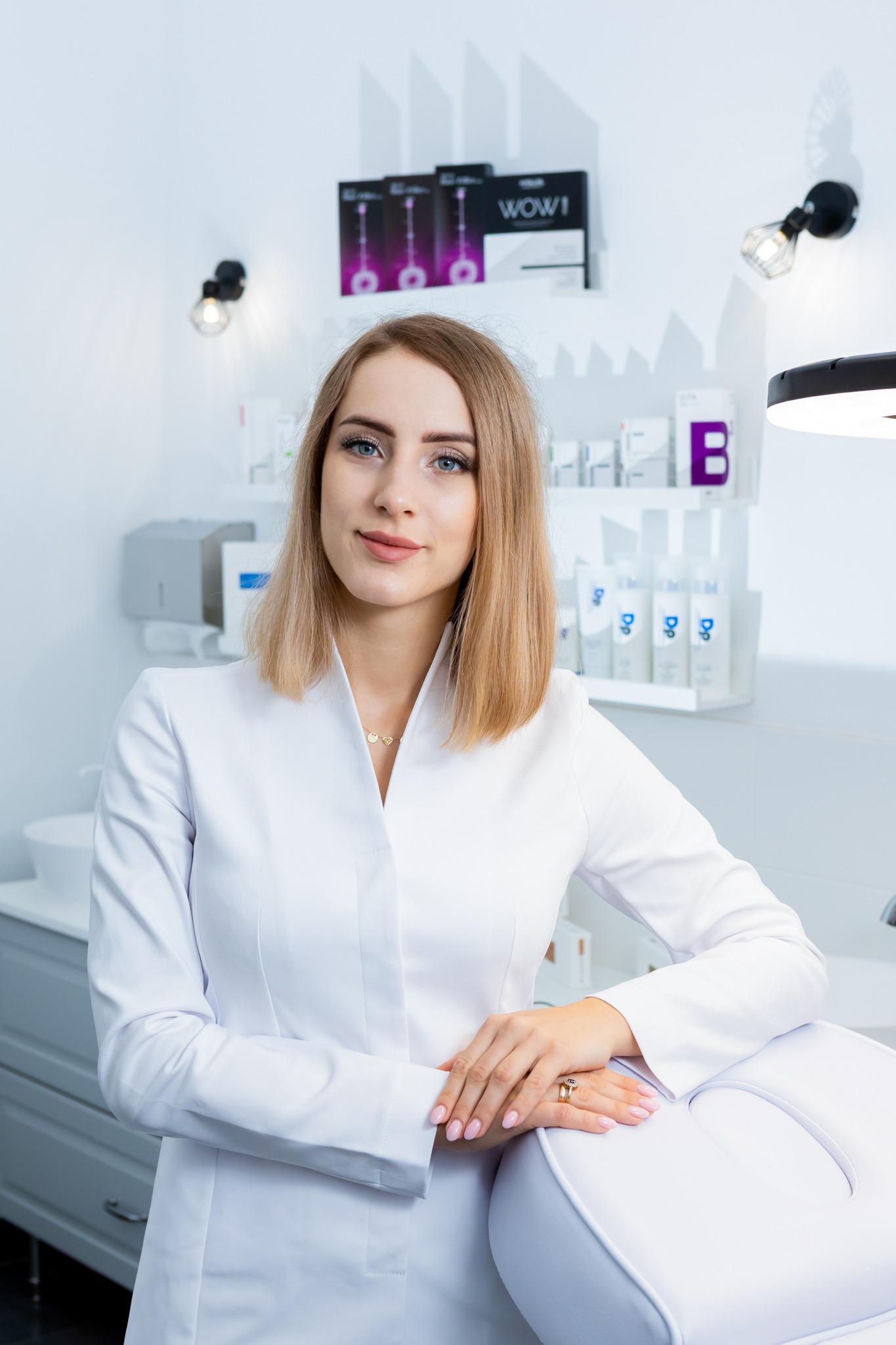 Portret kobiety w białym uniformie medycznym, opierającej się o biały fotel zabiegowy w sterylnym wnętrzu gabinetu kosmetycznego z półkami wypełnionymi kosmetykami i lampami punktowymi.