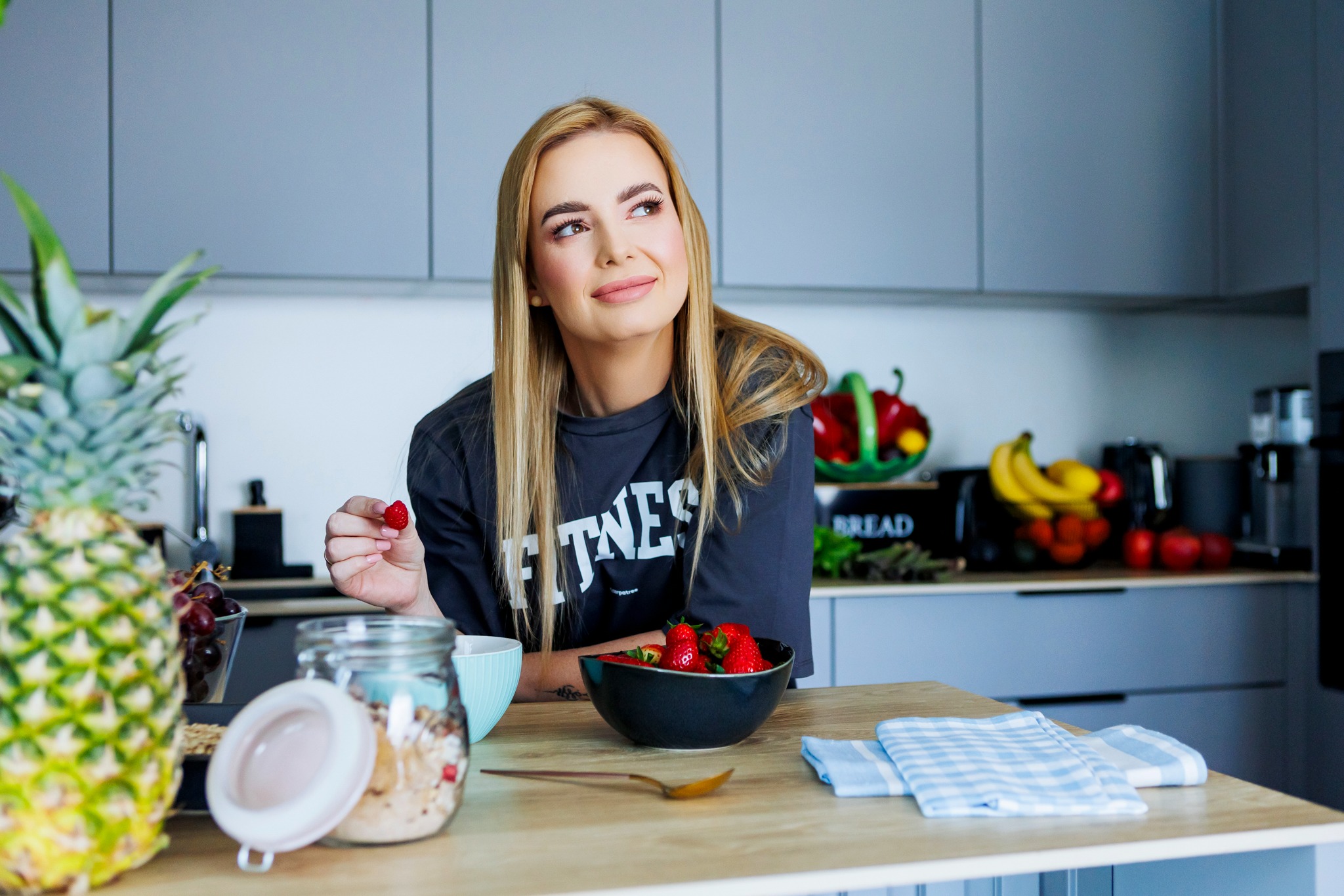 Kobieta w koszulce z napisem 'FITNESS' trzyma truskawkę w kuchni, obok ananas, miska truskawek, słoik z granolą i niebieska miseczka.