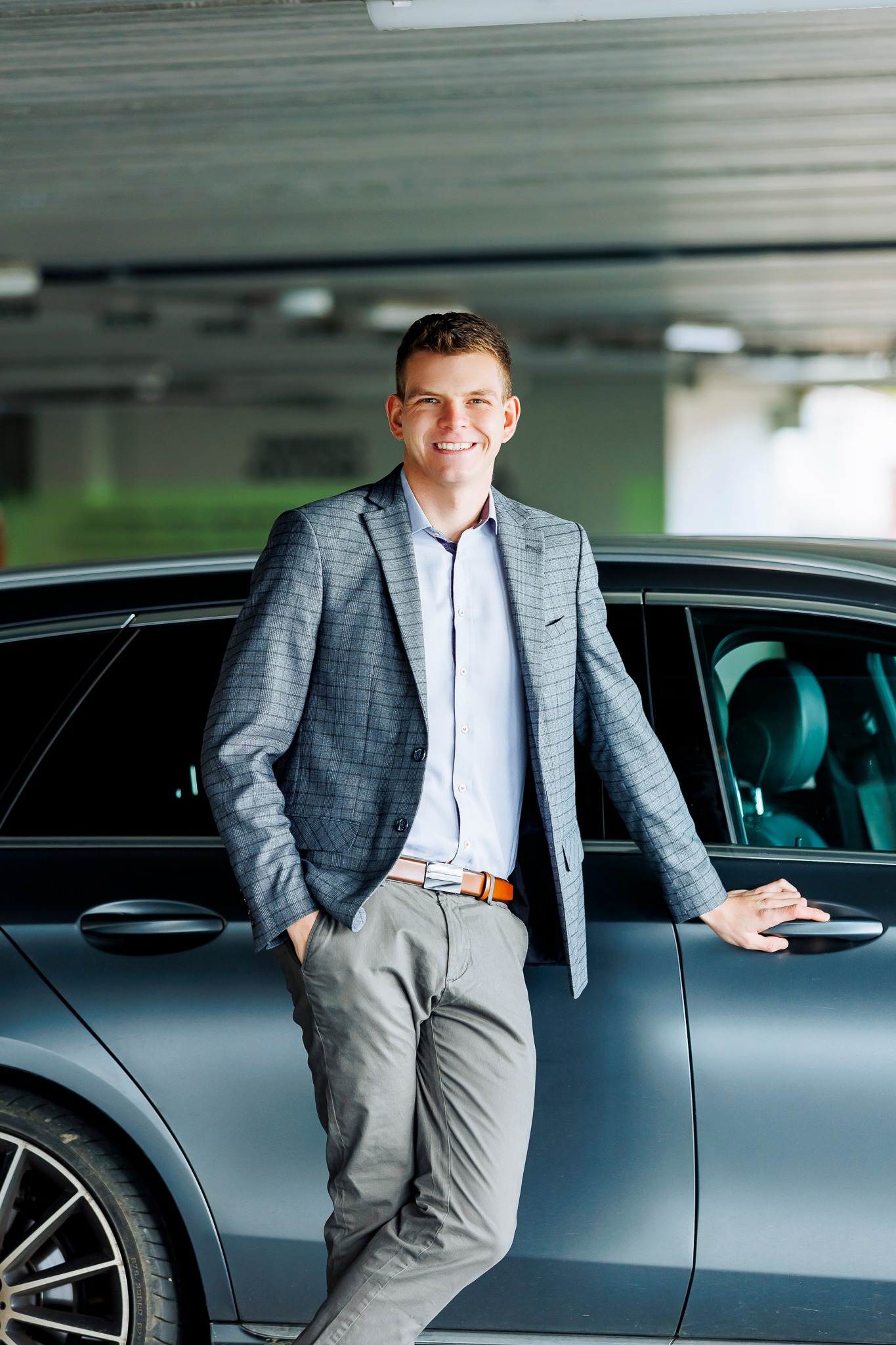 Uśmiechnięty mężczyzna w szarej marynarce w kratę i spodniach chino, oparty o ciemny samochód na tle parkingu podziemnego.