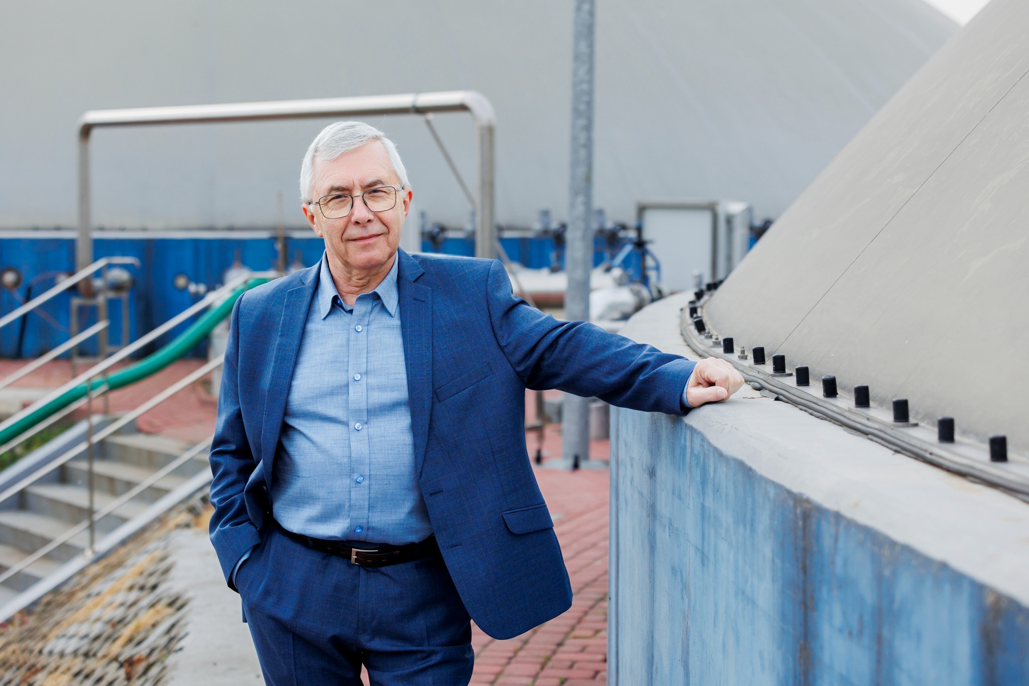 Mężczyzna w niebieskim garniturze opiera się o betonową konstrukcję przemysłową z widocznymi rurami i schodami w tle.