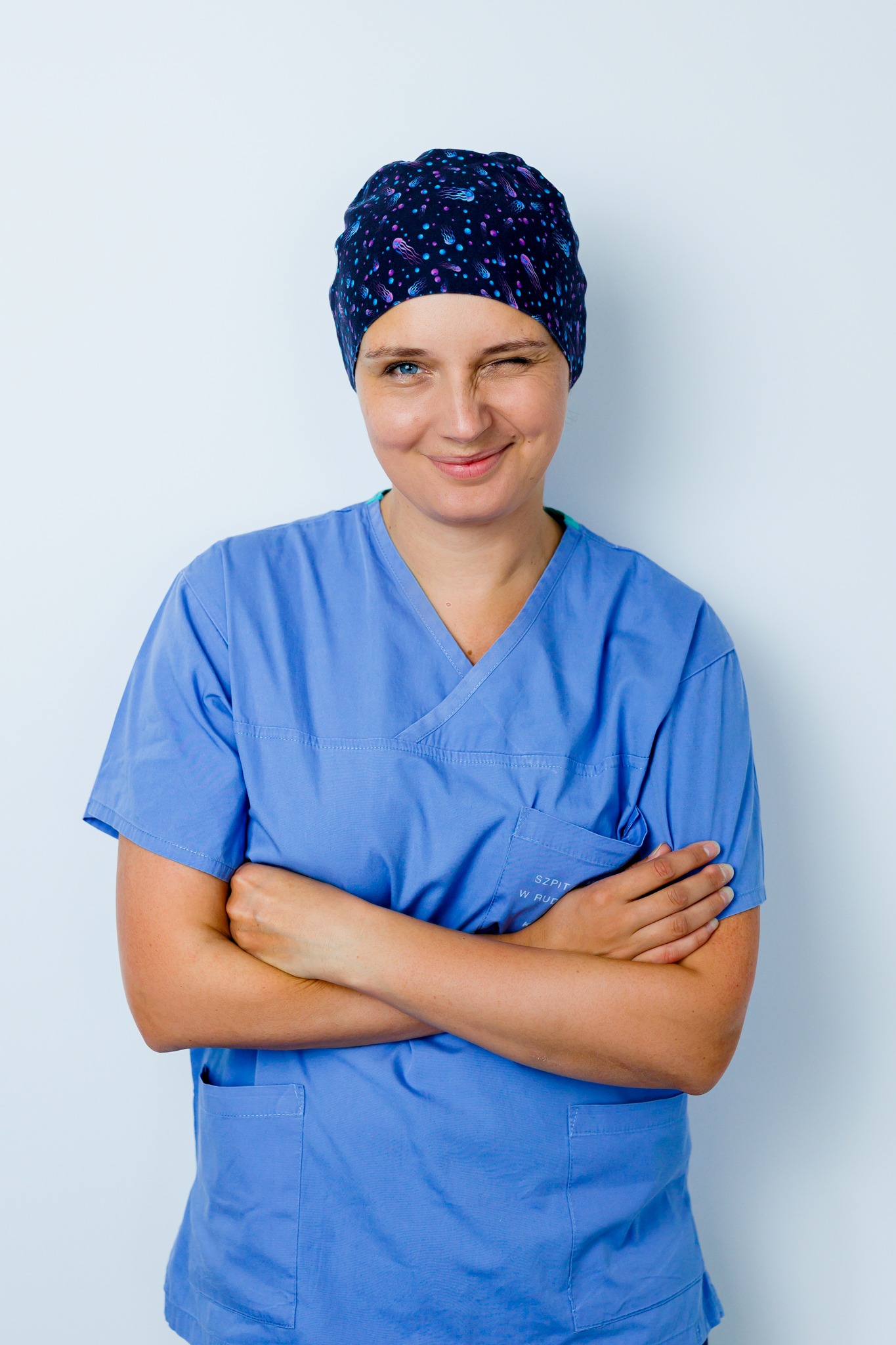 Uśmiechnięta kobieta w niebieskim uniformie medycznym i czepku we wzory, mruży jedno oko, pozując na białym tle.