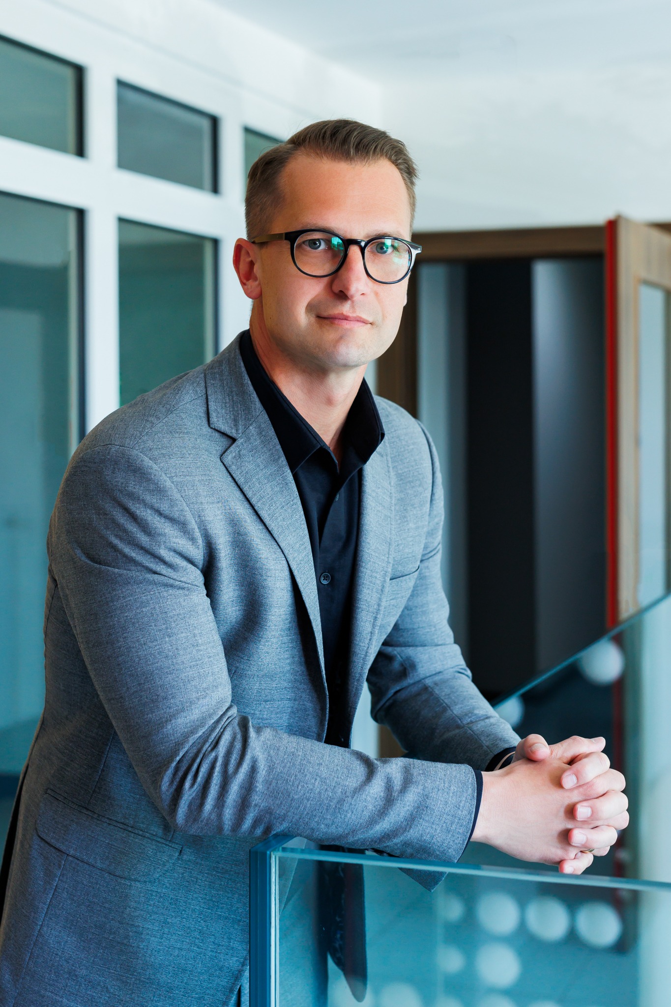 Mężczyzna w szarym garniturze i czarnej koszuli, oparty o szklaną balustradę, z rękami splecionymi przed sobą, spogląda w obiektyw.