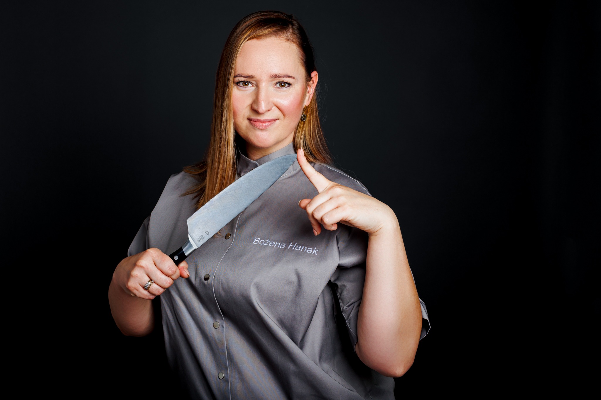 Portret kobiety w szarym uniformie kucharskim z wyhaftowanym imieniem 'Bożena Hanak', trzymającej duży nóż kuchenny, z uśmiechem spoglądającej w obiektyw na ciemnym tle.