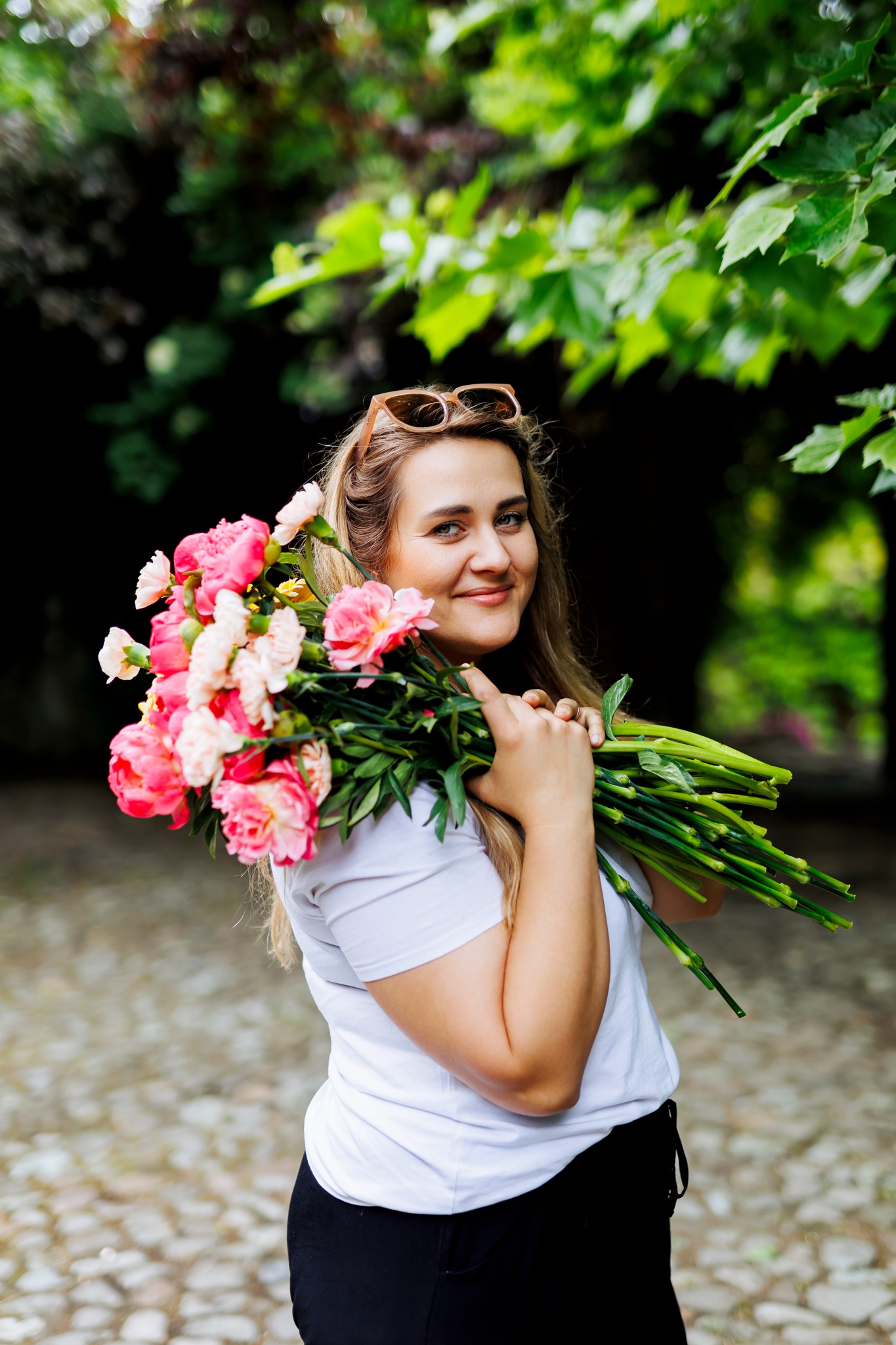 Uśmiechnięta blondynka w białej koszulce i czarnych spodniach trzyma na ramieniu duży bukiet różowych i białych piwonii na tle zieleni.