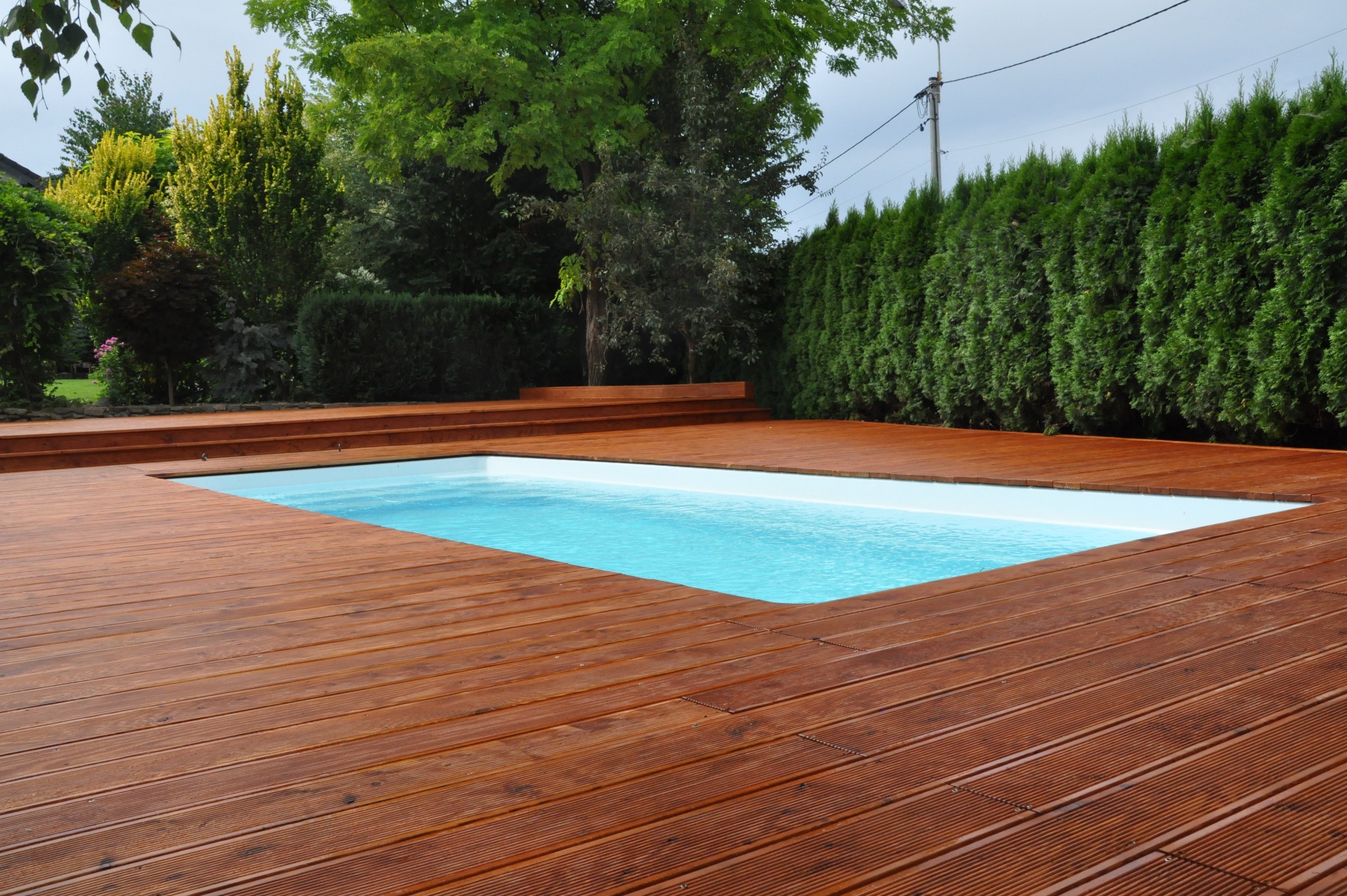 Nowoczesny basen otoczony tarasem z brązowych desek, z zielonym żywopłotem w tle i drzewami w oddali.