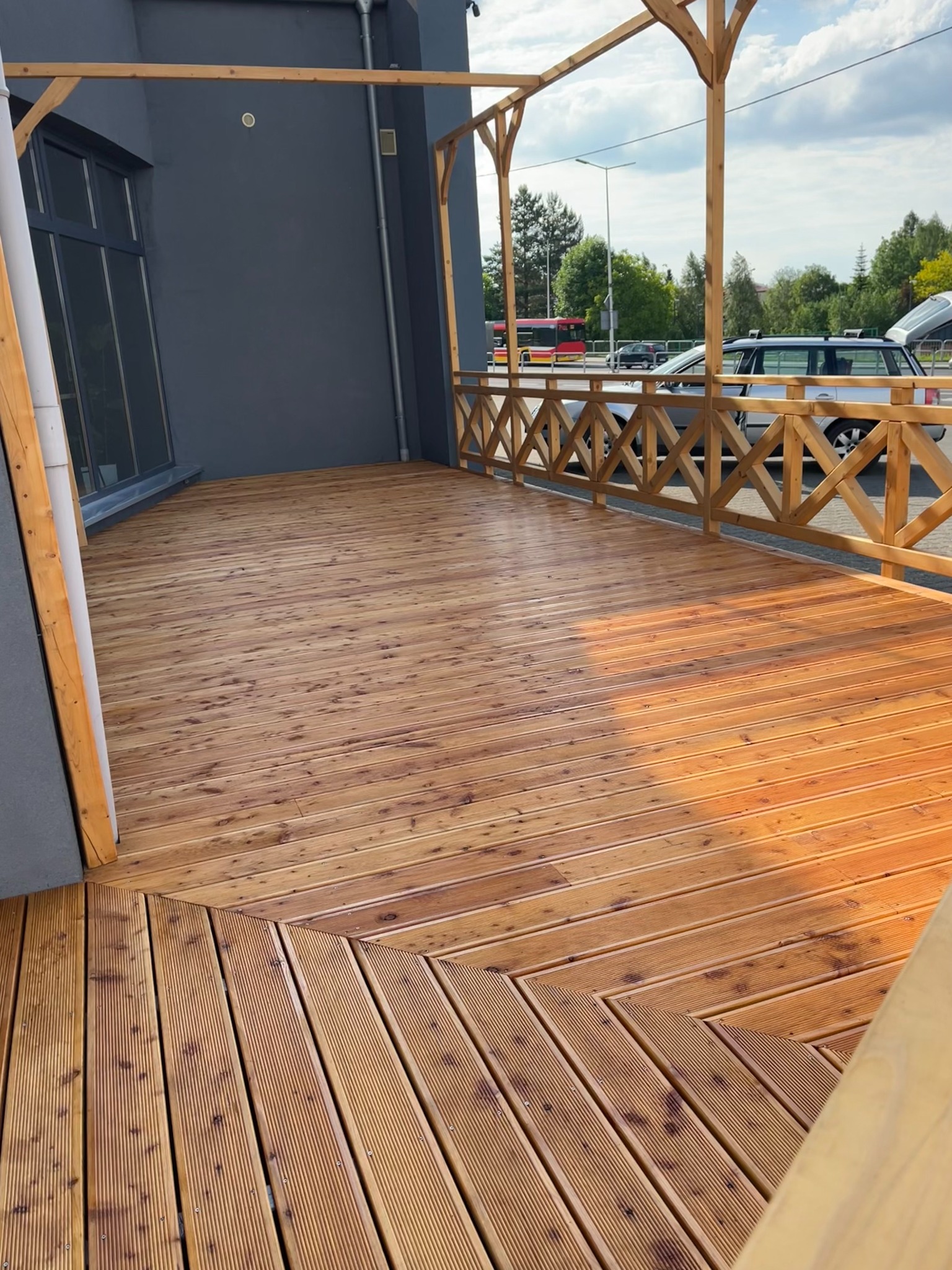 Nowoczesny taras drewniany z jasnego drewna, z geometrycznym wzorem ułożenia desek, otoczony drewnianą balustradą z motywem krzyży, na tle szarej elewacji budynku i zieleni drzew.