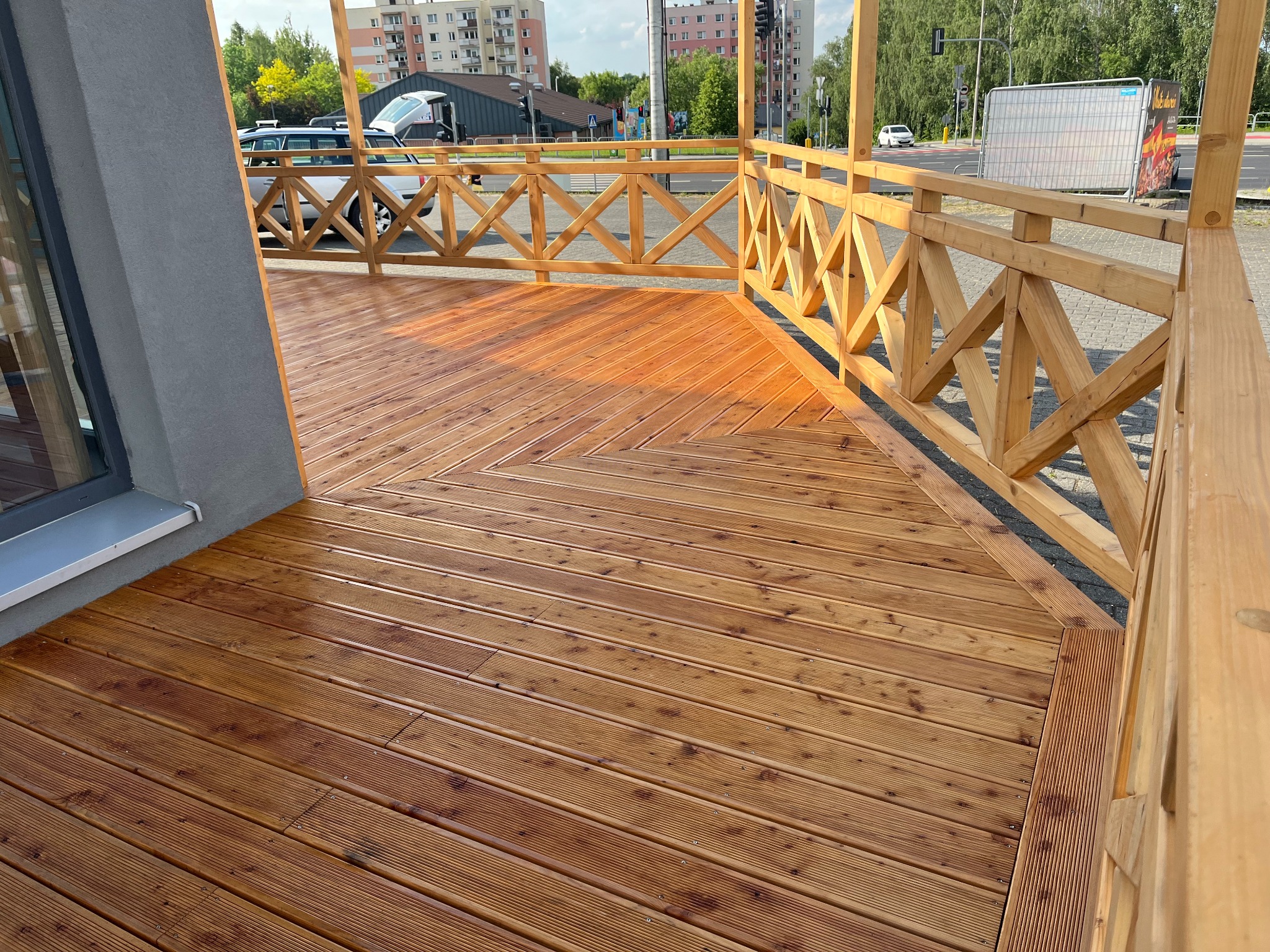 Świeżo wybudowany taras z drewnianych desek o ciepłym, brązowym odcieniu, z ozdobną, drewnianą balustradą w jasnym kolorze, na tle miejskiego krajobrazu z budynkami mieszkalnymi i ulicą.