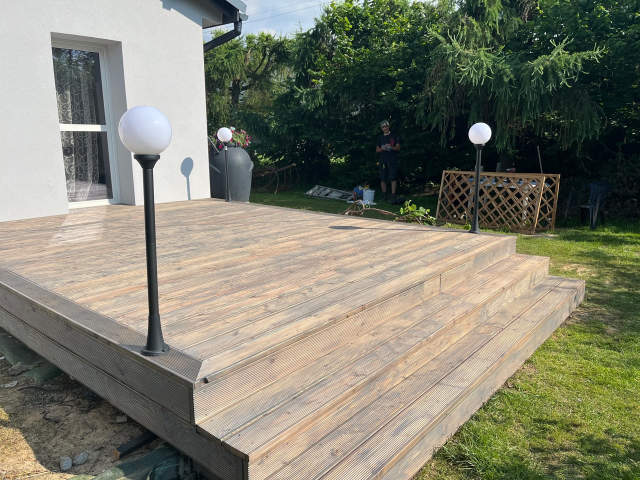 Drewniany taras z trzema stopniami i oświetleniem w postaci kulistych lamp, widok na ogród i częściowo na biały dom, w tle osoba oraz narzędzia sugerujące prace wykończeniowe.